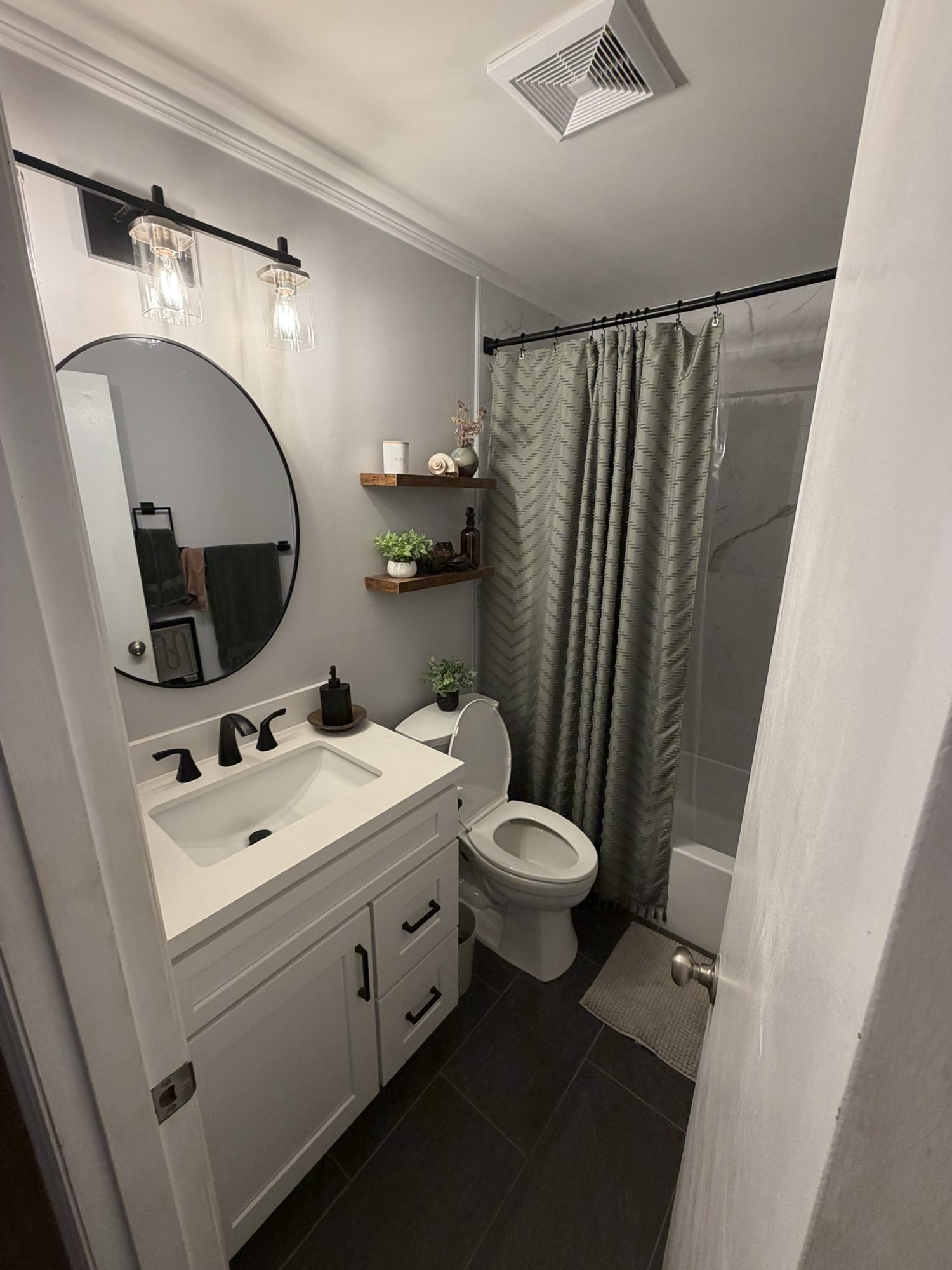 Bathroom with a white vanity, round mirror, floating shelves and dark tile floor